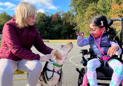  child in a wheelchair petting a seeing eye dog while its trainer looks on