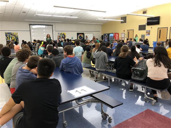 Students sitting in cafeteria listening to teacher.